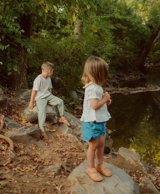 Two children exploring rocks by a creek wearing tan barefoot leather shoes and sandals